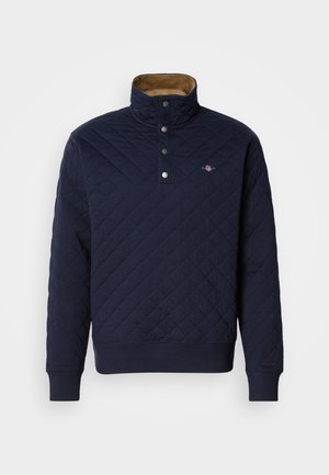 Marineblå quiltepullover med høy krage, fem-knappsglidelås og kordfløyelsforing. Har ribbestrikkede mansjetter og nederkant. Liten logodetalj på brystet.