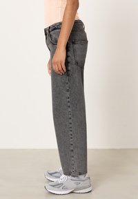 Persona tērpusies augsta vidukļa izbalējušās melnās džinsās un baltpelēkās sporta kurpēs, stāvot sāniski pret vienkāršu gaišu fonu.