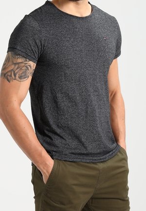 Mężczyzna w dopasowanej, ciemnoszarej koszulce z krótkim rękawem i oliwkowych spodniach, z rękami w kieszeniach, z tatuażem róży na lewej górnej części ramienia.