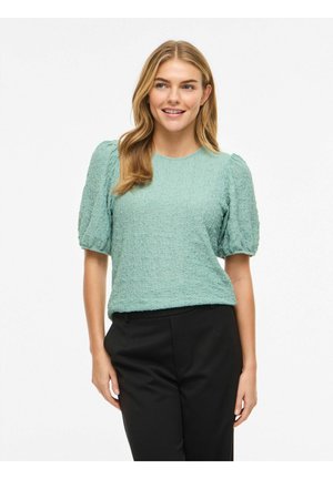 Hymyilevä nainen, jolla on pitkät vaaleat hiukset ja päällään teksturoitu vaaleanvihreä puffy-hihainen paita sekä mustat housut, seisoo yhdellä kädellä taskussa.