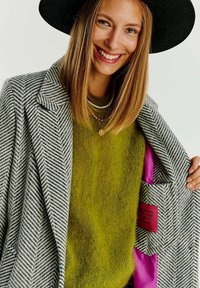 Šedý kabát ve vzoru herringbone s kontrastní růžovou podšívkou, kombinovaný se zeleným strukturovaným svetrem. Obsahuje kapsu s červeným detailem štítku.