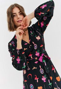 Czarna sukienka z różowymi, zielonymi i pomarańczowymi fantazyjnymi wzorami, z głębokim dekoltem w serek, długimi bufiastymi rękawami i talią z paskiem ozdobionym satynową kokardą.
