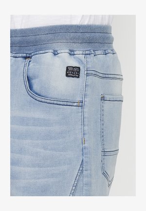 Svijetloplave traper jogger hlače s elastičnim pojasom, koje imaju bočne džepove i malu crnu oznaku brenda. Glatka tekstura s detaljima šivanja.