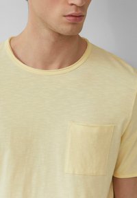 Geel T-shirt met korte mouwen, gemaakt van een zachte, gestructureerde stof. Heeft een ronde halslijn en een enkele patch pocket op de linkerborst.