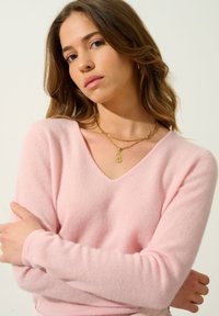 Hellrosa Cashmere-Pullover mit V-Ausschnitt, langen Ärmeln und weicher Textur. Übereinander getragene goldene Halsketten verleihen einen dezenten Glanz.