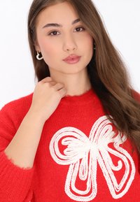 Rode gebreide trui met een groot wit strikontwerp. Het materiaal lijkt textuur te hebben, met een licht versleten kraag en manchetten.