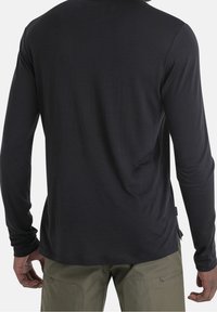 Mann in einem enganliegenden schwarzen Langarmshirt und olivgrünen Hosen, von hinten gezeigt mit entspannten Armen an den Seiten.