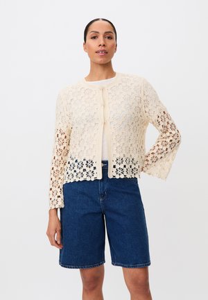 Frau trägt eine cremefarbene, florale Häkeljacke über einem weißen Top und dunkelblaue, knielange Jeansshorts, steht mit einer Hand in der Hüfte.