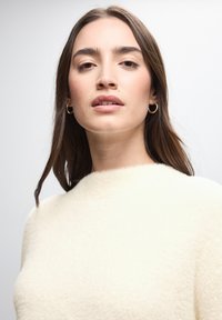 Cremefarbener Strickpullover mit einer weichen, flauschigen Textur und einem runden Ausschnitt, kombiniert mit goldenen Creolen. Glatter, heller Hintergrund.