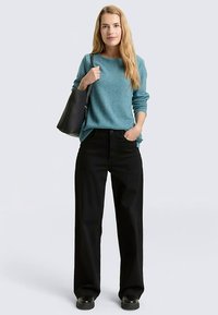 Pull en tricot bleu clair, coupe ample, associé à un pantalon noir à jambes larges taille haute. Le mannequin tient un sac à main noir. Texture lisse, style décontracté.