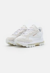 Ett par vita och ljusbeige Reebok Classic-sneakers med texturerade sulor och snörningsdesign på en vit bakgrund.