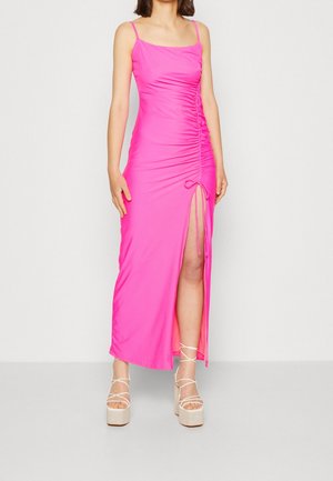 Mujer con un vestido fruncido de color rosa brillante con una abertura en el muslo y tacones de plataforma blancos con tiras, posando frente a un fondo liso.