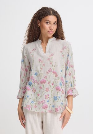 Kvinde iført en lysebluse med lyserøde og blå blomsterprint og kvartærmer, kombineret med lyse bukser.