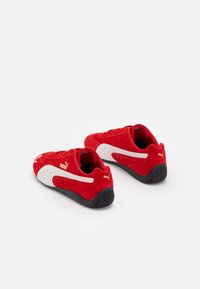 Paire de baskets Puma rouges avec des bandes blanches et des semelles noires, photographiée de dos sur un fond blanc uni.