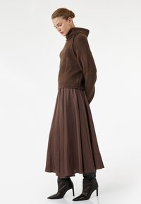 Brązowy sweter dziany z wysokim kołnierzem, zestawiony z luźną, długą brązową spódnicą i czarnymi szpilkami z ostrym noskiem. Ogólnie gładka tekstura.