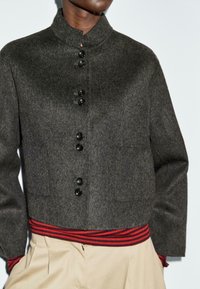 Giacca di lana grigia scura con bottoni e colletto alto, tasca a pattina, orlo a strisce rosse e nere, indossata con pantaloni beige e mani nelle tasche.
