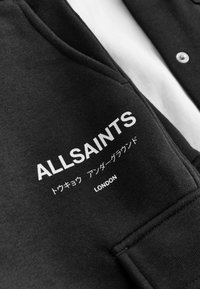 Čierna mikina s bielym textom "ALLSAINTS" a "LONDON", so hladkou textúrou a gombíkmi pozdĺž prednej časti.