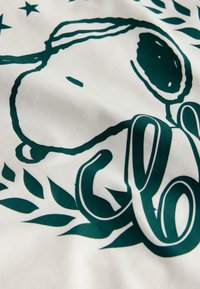 Weißer Stoff mit einer grünen Illustration eines Cartoon-Hundes und dem Buchstaben "c", umgeben von Blättern und Sternen.