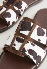 Braune Rutsch-Sandalen mit Kuhflecken, die weiße und braune Texturen aufweisen. Zwei breite Riemen überqueren eine braune Sohle. Auffällige Hardware-Akzente.