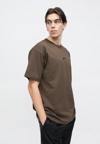 Νεαρός με κοντά σκούρα μαλλιά, ντυμένος με καφέ μπλουζάκι Nike και μαύρο παντελόνι, με τα χέρια στις τσέπες, που κοιτάζει πλάγια πάνω σε απλό ανοιχτόχρωμο φόντο.