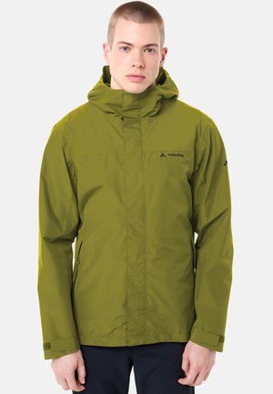 Giovane uomo che indossa una giacca impermeabile con cappuccio verde oliva con il logo VAUDE, in piedi davanti a uno sfondo chiaro e uniforme.