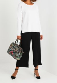 Pantalones anchos negros, camisa blanca de manga larga y un bolso con estampado de camuflaje y detalles florales. Tacones de aguja negros completan el conjunto.