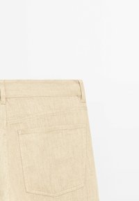 Pantalon en denim beige montrant la poche arrière, les passants de ceinture et le tissu texturé sur fond blanc.