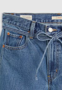 Calças de ganga azuis com corte reto, costura visível, cintura amarrada e bolsos frontais com detalhes em cobre e fecho de botão.