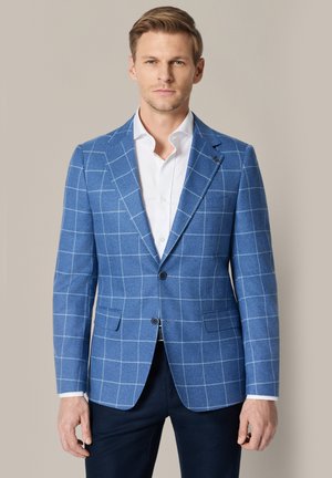 Mężczyzna ubrany w niebieską marynarkę w kratę windowpane, białą koszulę i ciemne spodnie, stojący na tle jednolitego beżowego tła.