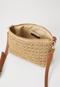 Vävt beige bagage med en texturerad yta och en guldfärgad dragkedja. Utrustad med ett slätt beige axelband och en beige tyginsida.