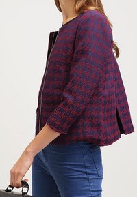 Textured wool blend jacket in dark purple with red geometric patterns, zip front, and three-quarter sleeves; features a split back design.