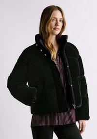 Melna pūkaina virsjaka ar spīdīgu apdari, augstu apkakli, divām priekšējām kabatām un aizdari ar spiedpogām, valkāta virs svītraina topa.