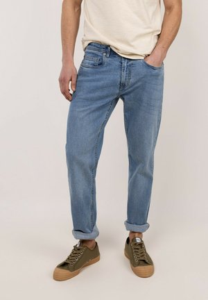 Hombre con vaqueros azul claro remangados, camiseta beige y zapatillas verde oliva, de pie con la mano en el bolsillo.