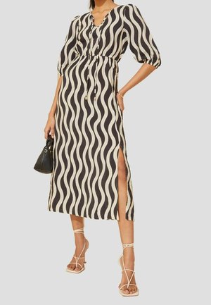 Femme portant une robe midi noire et beige à motif ondulé avec une fente latérale, des manches bouffantes, tenant un sac à main noir, et portant des talons beiges à brides.