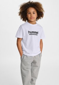 Vit bomullströja med "hummel SPORTSWEAR" tryckt i svart, kombinerad med grå träningsoverallsbyxor, som visar en avslappnad passform och rundad halsringning.