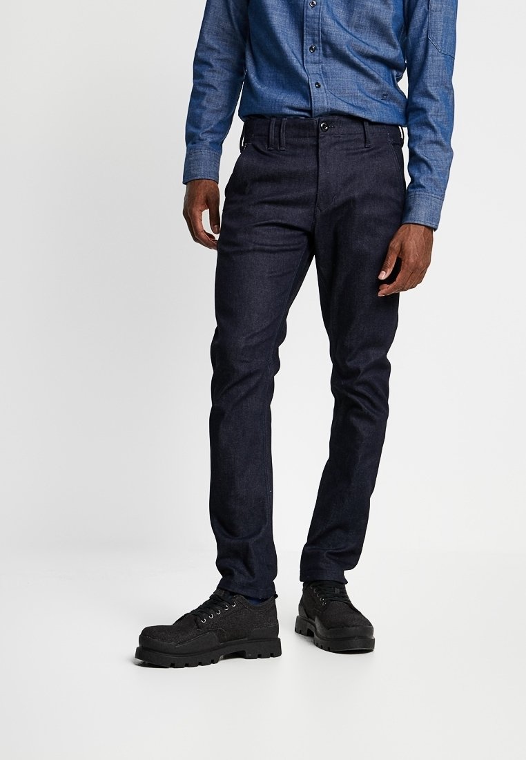 Dark blue denim trousers with a slim fit, featuring a classic waistband and front pockets, paired with black textured sneakers.