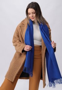 Manteau en fausse fourrure marron, écharpe bleue avec bords fringés, haut crème et pantalon beige. Textures douces, coupe décontractée et contraste de couleurs vives.