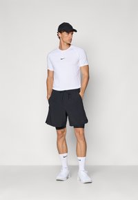 Weißes Sportshirt mit schwarzem Nike-Logo, kombiniert mit schwarzen Shorts und weißen Turnschuhen, vor neutralem Hintergrund stehend.