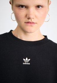 Juodas džemperis su raukšlėtu apykakle ir balta siuvinėta Adidas logotipu krūtinėje. Lygus paviršius, laisvalaikio stilius, standartinis šiupinys.