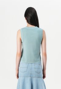 En kvinne med langt mørkt hår iført en lyseblå ermeløs topp og et denimskjørt, vist fra baksiden mot en enkel hvit bakgrunn.