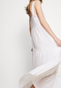 Hvid, flydende kjole lavet af letvægtsstof, med blonder detaljer og en svajet silhuet, fremhævet af lagdelt tekstur og detaljer.