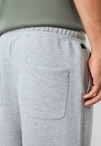 Nahaufnahme einer Person, die hellgraue Jogginghosen mit elastischem Bund und einer Rückentasche trägt, mit der Hand an der Seite, während sie ein olivgrünes Oberteil anhat.
