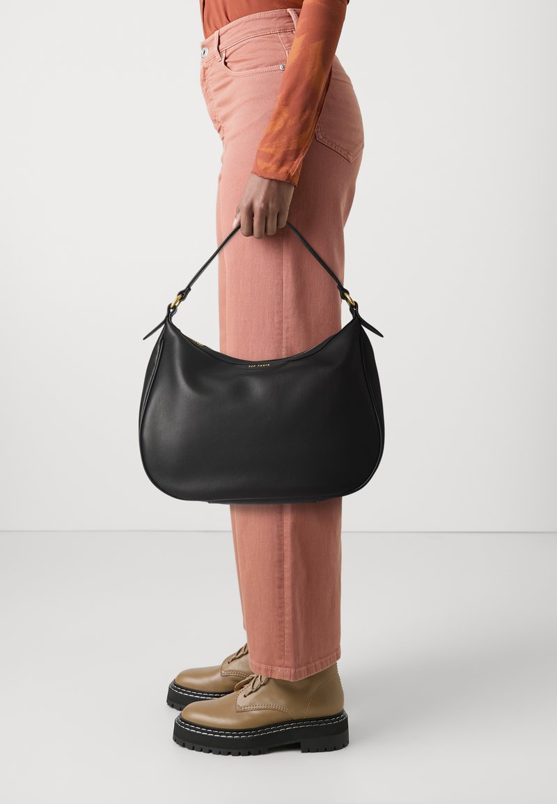 Sac de épaule en cuir noir avec une forme courbée, présentant des accents en métal doré. Porté par une personne en pantalon rose et chaussures beiges.