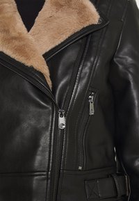 Zwarte leren jas met een nepbontkraag, diagonale ritssluiting aan de voorkant en ritssluitingen aan de zijkanten. Voorzien van een riem en een gestructureerde afwerking voor extra detail.