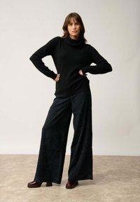 Schwarzer Rollkragenpullover kombiniert mit weiten, strukturierten dunkelgrauen Hosen. Das Outfit wird durch klobige bordeauxfarbene Schuhe ergänzt.