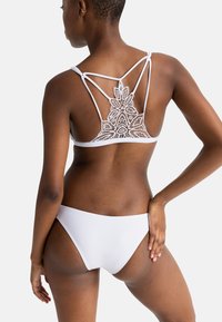 Baltas bikini komplektas, kurį sudaro nėriniuotas liemenėlė su sudėtingais gėlių raštais ir plonomis petnešėlėmis, derinamas su atitinkamomis žemomis kelnaitėmis.