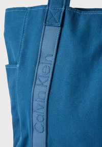 Μπλε καμβάς τσάντα ώμου Calvin Klein με ραμμένη λαβή και πλαϊνή τσέπη, που δείχνει τη λεπτομερή υφή του υφάσματος και το λευκό ράψιμο.