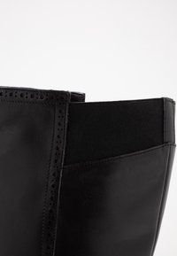 Bota de tobillo de cuero negro con acabado texturizado, panel elástico y perforaciones decorativas a lo largo de la costura. Diseño elegante y minimalista.