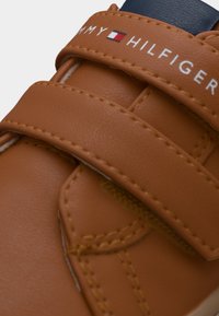Καφέ συνθετικά παπούτσια με δύο λουράκια βέλκρο, με λευκή επωνυμία "Tommy Hilfiger" και αντιθετικές σκούρες μπλε λεπτομέρειες.