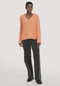 Frau stehend in einem orangefarbenen Strick-V-Ausschnitt-Pullover, dunklen grünen Cordhosen und dunkelbraunen Loafers vor einem schlichten hellen Hintergrund.